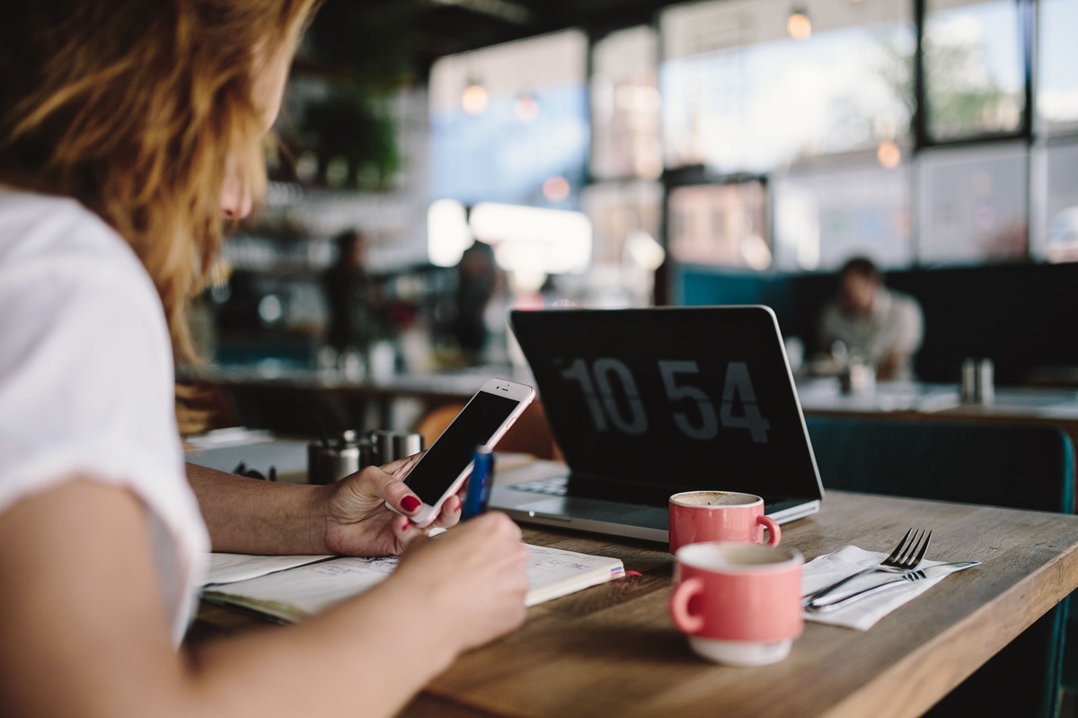 Woman holding mobile phone in coffee shop with laptop